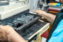 Cultivo irreconocible anciano artesano en delantal y anteojos preparando marco metálico para impresión de tipografía en estudio - foto de stock