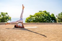 Side view of flexible female in activewear performing Eka Pada Sarvangasana on mat on dry ground during yoga session in park against green trees and cloudless blue sky in sunlight — Stock Photo
