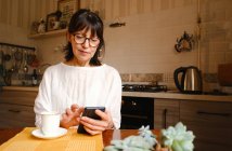 Mujer satisfecha en anteojos mensajes de texto en el teléfono inteligente mientras está sentado en la mesa con una taza de café en la mesa en la cocina - foto de stock