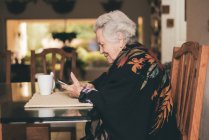 Вид збоку усміхненої літньої жінки в теплому одязі, що сидить за столом з таблеткою і чашкою чаю, дивлячись на екран — стокове фото