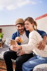 Giovane donna sorridente con un bicchiere d'acqua seduta vicino al fidanzato barbuto e scattare selfie con smartphone sul divano in terrazza — Foto stock