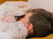 Vista lateral del niño enfermo con gripe acostado con los ojos cerrados debajo de la manta en el sofá y durmiendo en la sala de estar en casa - foto de stock
