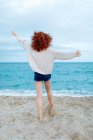 Back view full length of unrecognizable barefoot female traveler standing on sandy coast washed by foamy waves of blue sea — Stock Photo
