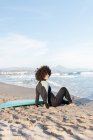Vista laterale corpo completo di surfista donna scalza in muta seduta sulla spiaggia sabbiosa vicino al mare ondulato — Foto stock