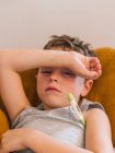 De cima de menino doente medir a temperatura com termômetro eletrônico enquanto deitado no sofá em casa e com gripe — Fotografia de Stock