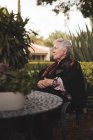 Seitenansicht einer nachdenklichen älteren Frau mit grauen Haaren, die kariert am Gartentisch sitzt und an sonnigen Tagen wegschaut — Stockfoto