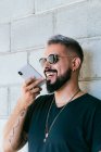 Glücklicher bärtiger Typ mit Tätowierungen in schwarzem T-Shirt und Sonnenbrille, der in der Nähe einer Hauswand steht und bei Tageslicht eine Audiobotschaft mit dem Smartphone aufzeichnet — Stockfoto