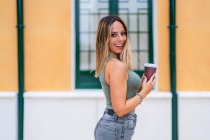Mujer positiva con taza de papel de café para llevar sonriendo y mirando a la cámara mientras está de pie cerca del edificio en la calle de la ciudad - foto de stock