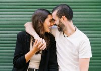 Alegre apaixonado jovem casal hispânico em roupas casuais rindo com os olhos fechados, enquanto abraçando e tocando as testas perto de parede verde na rua da cidade — Fotografia de Stock