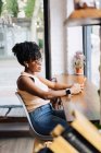 Vista laterale di fiduciosa giovane afro-americana millenaria con capelli ricci scuri in abito alla moda e occhiali da sole seduti a tavola in caffè moderno e messaggistica su smartphone — Foto stock