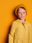 Petit garçon sans émotion en imperméable à la mode et bonnet chapeau debout regardant loin sur fond jaune en studio — Photo de stock