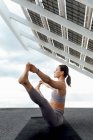 Vue latérale complète du corps de la femme sportive en vêtements de sport pratiquant la posture Ubhaya Padangushthasana près du panneau photovoltaïque moderne pendant l'entraînement de yoga dans la rue — Photo de stock