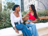 Serias amigas afroamericanas hablando y mirándose mientras están sentadas en el banco cerca de plantas verdes en la calle con edificios - foto de stock