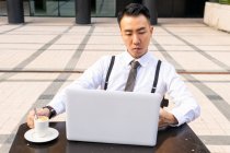 Schnodderiger junger asiatischer Unternehmer mit Tasse Heißgetränk und Netbook, der bei Tageslicht auf den Bildschirm in der städtischen Cafeteria blickt — Stockfoto