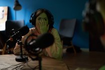 Jeune animatrice radio en vêtements décontractés et écouteurs assis à table avec microphone et communiquant avec un collègue anonyme lors de l'enregistrement de podcast en studio — Photo de stock