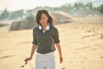 Side view of peaceful thoughtful Asian female looking down listening to song from wireless headphones while walking on sandy shore — Stock Photo