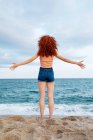 Vista trasera de longitud completa de viajera femenina descalza irreconocible de pie en la costa arenosa bañada por olas espumosas de mar azul - foto de stock