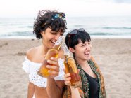Люблячи багатоетнічну пару жінок, які чіпляються за пляшки пива і насолоджуються літнім днем на березі моря — стокове фото