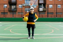 Положительная зрелая женщина в спортивной одежде и повязке смотрит в камеру, стоя с мячом во время игры в баскетбол — стоковое фото