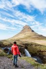 Vista posteriore del viaggiatore irriconoscibile in tuta capispalla in piedi con macchina fotografica vicino alla cascata e ammirando uno spettacolare scenario di montagna a Kirkjufell in Islanda durante il giorno — Foto stock