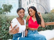 Deliziose amiche afroamericane che si autoritrattano sul cellulare mentre siedono sulla panchina vicino a piante verdi in strada con edifici moderni — Foto stock