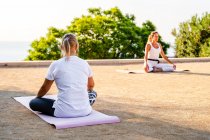 Ladies practicing lotus position together while sitting on mats on sandy terrace in sunny day — Stock Photo