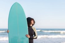Side view of young thoughtful female surfer in wetsuit with surfboard standing looking away on seashore washed by waving sea — Stock Photo
