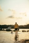 Ganzkörper eines anonymen männlichen Besitzers mit Stiefeln in den Händen, der an einem Sommertag in der Natur in der Nähe eines laufenden Hundes im Wasser spazieren geht — Stockfoto