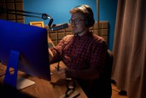De cima focado jovem macho em camisa quadriculada e óculos usando computador e falando em microfone durante a gravação de podcast no estúdio escuro — Fotografia de Stock