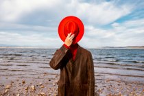Неузнаваемый человек в красной шляпе и пальто, стоящих на берегу с дроблеными камнями летом — стоковое фото