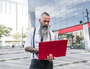Hipster trabajador masculino en desgaste formal netbook de navegación mientras está de pie en la calle urbana - foto de stock