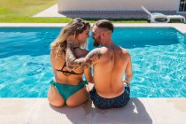 Visão traseira de casal amoroso em trajes de banho olhando uns para os outros enquanto acaricia na borda da piscina em resort tropical — Fotografia de Stock