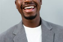 Cultivar empresário masculino afro-americano em terno formal sorrindo amplamente enquanto está de pé contra fundo cinza e olhando para a câmera — Fotografia de Stock