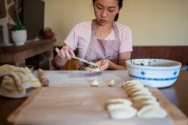 Frau in lässiger Kleidung und Schürze, die Knödel mit Fleisch füllt, während sie traditionelle chinesische Jiaozi in der Küche zubereitet — Stockfoto