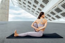 Vue arrière du corps complet de la femme sportive méconnaissable en vêtements de sport assis sur le tapis tout en pratiquant le yoga asana avec torsion près du panneau solaire sur la rue — Photo de stock