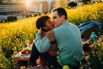 Jovem senhora afro-americana com cabelo encaracolado abraçando e beijando bochecha de namorado romântico com olhos fechados durante piquenique no prado florescendo no dia ensolarado — Fotografia de Stock