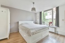 Interno di camera da letto arredata con letto rivestito con copriletto bianco posto vicino armadio in legno sotto lampada — Foto stock