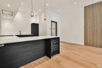 Интерьер просторной кухни с минималистской черной мебелью и мини-холодильником в современной квартире с белыми стенами и деревянным паркетным полом — стоковое фото