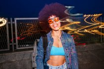 Весела жінка з сонцезахисними окулярами дивиться на камеру, стоячи біля паркану зі світловим ефектом у вечірній час — стокове фото