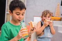 Веселі діти сміються і їдять свіжі бутерброди, сидячи за столом і п'ючи сік у світлій кімнаті вдома — стокове фото