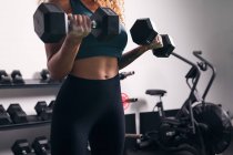 Культивировать сильную спортсменку с вьющимися волосами делая упражнения на бицепсах с гантелями во время тренировки в тренажерном зале — стоковое фото