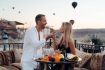 Pareja tomados de la mano y mirándose mientras disfrutan de una cena romántica y se alimentan mutuamente con frutas en la terraza con globos de aire caliente en el fondo en Capadocia - foto de stock