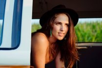 Симпатичная брюнетка в шляпе в винтажном фургоне и сидит на сиденье в солнечный день — стоковое фото