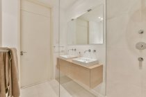 Белые раковины на стене с зеркалом, расположенным рядом со стеклянной душевой кабиной в светлой просторной ванной комнате с инструментами и яркой подсветкой — стоковое фото