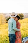 Visão lateral da jovem mulher afro-americana feliz com cabelo escuro curto sorrindo enquanto abraça o namorado anônimo com os olhos fechados em pé no prado florido no dia ensolarado — Fotografia de Stock