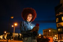 Femme positive avec une coiffure afro et des vêtements modernes messagerie texte sur téléphone portable tout en se tenant debout dans la rue avec des bâtiments et des lampadaires en soirée — Photo de stock