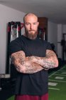 Forte sportivo adulto con barba e tatuaggi sulle braccia incrociate guardando la fotocamera in palestra — Foto stock