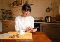Mujer satisfecha en anteojos que envían mensajes de texto en el teléfono inteligente mientras está sentada en la mesa con una taza de café colocada cerca de bellotas en la mesa en la cocina - foto de stock