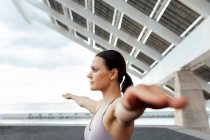 Визначена жінка в активному одязі з розтягнутими руками займається вправами йоги на вулиці біля сучасної фотоелектричної панелі під час тренувань у місті — стокове фото