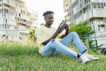 Cuerpo completo de joven hombre negro sonriente con camiseta de color amarillo claro y vaqueros azules y zapatillas de deporte sentados en la hierba y el teléfono inteligente de navegación - foto de stock